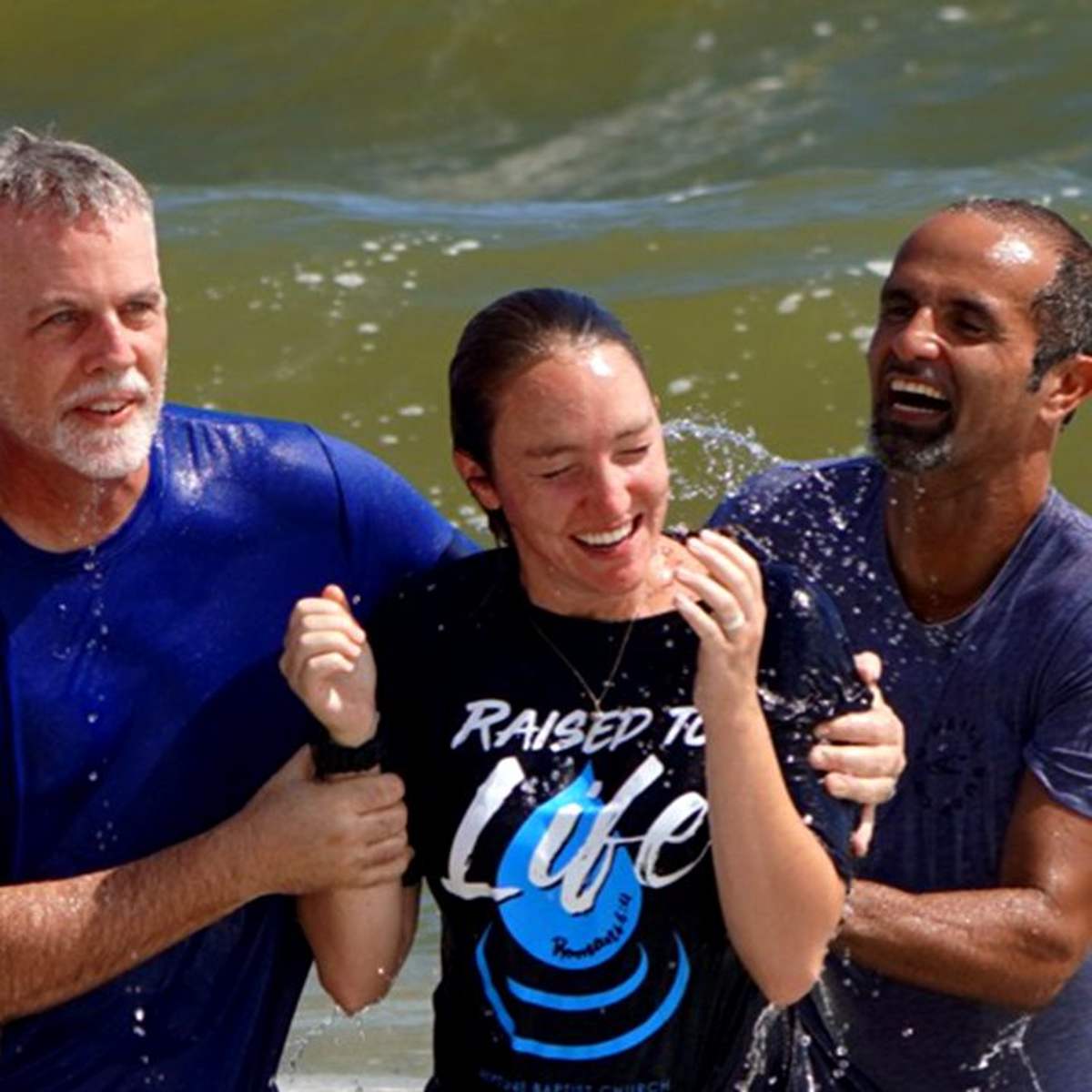 A newly baptized young woman smiling in the surf with two pastors